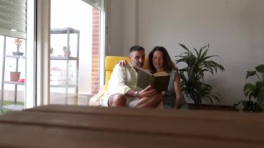 Bir erkek ve bir kadın, pencerenin kenarındaki rahat bir sandalyede oturmuş, elindeki kitaba dalmış birlikte sakin bir anın tadını çıkarıyorlar.