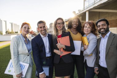 Şehirdeki çeşitli iş adamları, ellerinde tablet ve klasörler tutarak, güven ve başarı yayarak mutlu bir şekilde iş birliği yapıyorlar. Gökdelenli kentsel ortam profesyonel atmosfere katkı sağlıyor