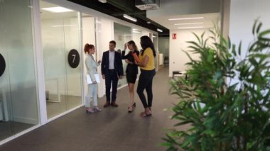 Dört profesyonel iş adamı modern bir ofis koridorunda sohbet ediyorlar. Atmosfer işbirlikçi ve ilgi çekici.