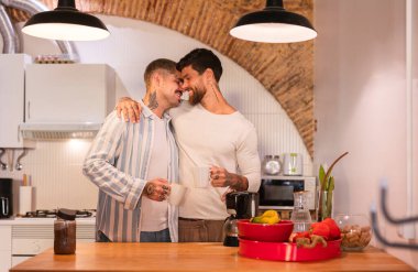 İki adam rahat bir mutfakta kahve fincanlarını kucaklıyor, gülümsüyor ve mutlu bir şekilde modern bir iç mekanda, tuğla bir duvar ve sıcak bir aydınlatma ile, samimi bir aşk ve birliktelik sahnesi yaratıyor.