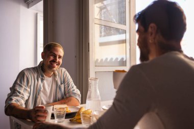 İki adam bir masada oturmuş, kahvaltı ediyor ve sohbet ediyorlar. Hayatlarının mutlu ve samimi bir anını gösteriyorlar.