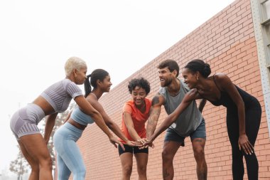 Spor kıyafetleri giyen beş gülümseyen sporcu, şehirde tuğla duvarın yanında antrenman yapmadan önce el ele tutuşuyor, takım çalışması ve motivasyon gösteriyorlar.