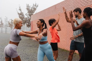 Bir grup çok ırklı sporcu, sisli bir şehir ortamında yaptıkları başarılı bir çalışma sonrasında başarılarını ve yoldaşlıklarını kucaklayarak beşlik çakıyor.