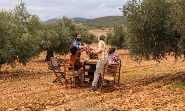 Çoklu etnik gruptaki çiftçiler, zeytin bahçesindeki ahşap masada oturmuş, başarılı bir hasat kutlamaları yaparken içki içiyorlar.