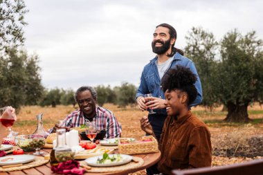 Çok ırklı bir grup çiftçi birlikte hoş bir öğle yemeği molası veriyor, zeytin koruluğunun ortasında bir ahşap masada oturup, yiyecek, içecek ve kahkaha paylaşıyorlar.