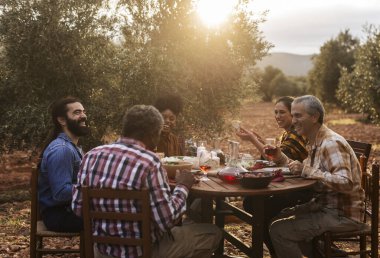 Çok ırklı bir grup çiftçi birlikte zeytinlikte ahşap bir masada yemek yiyorlar, hasatı yiyecek ve içeceklerle kutluyorlar, gün batımının ve birbirlerinin arkadaşlığının tadını çıkarıyorlar.