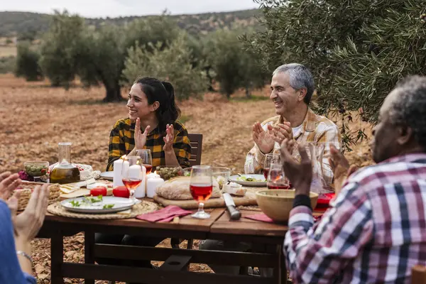 Çok ırklı ve çok nesilli çiftçiler zeytin koruluğunda bir masada kutlama yemeği yiyor ve alkışlıyorlar.