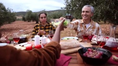 Çok ırklı bir grup çiftçi, zeytin bahçesindeki ahşap bir masada sağlıklı bir öğle yemeği yiyorlar. Başarılı hasatı gülümseme ve konuşmalarla kutluyorlar.