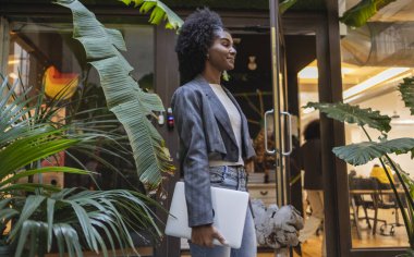 Afro saç stili dizüstü bilgisayarı olan kendine güvenen genç yönetici, yemyeşil bitkilerle süslenmiş modern çalışma odasına giriyor, iş yerinde çeşitliliği ve sosyal içeriği somutlaştırıyor.