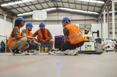 Mühendislerden oluşan çok ırklı bir ekip bir robot aracın etrafında çömelmiş, performansını analiz ediyor ve teknolojik olarak gelişmiş bir fabrikadaki potansiyel gelişmeleri tartışıyorlar.