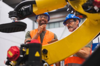 Güvenlik yelekleri ve başlık takan iki çok ırklı mühendis fabrika ortamında bir robot kolu tartışıyor ve analiz ediyor. Endüstriyel otomasyonda takım çalışması ve yenilik sergiliyorlar.