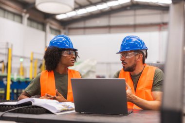 Güvenlik gözlüğü ve şapka takan iki endüstri mühendisi büyük bir fabrikada dizüstü bilgisayara bakarken bir projeyi tartışıyorlar.