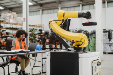 African american female engineer multitasking with a laptop and phone while programming a robotic arm in an industrial factory clipart