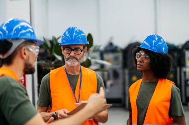Çağdaş bir fabrikada bir projeyi tartışan güvenlik yelekleri ve başlıklar giyen çok ırklı mühendisler takım çalışması, uzmanlık ve endüstriyel işbirliği.