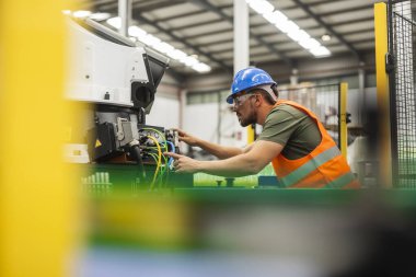 Focused industrial engineer wearing safety vest and hardhat operating robotic machinery in a modern factory, highlighting advanced manufacturing processes and skilled workforce clipart