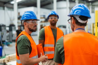 Güvenlik yelekleri ve miğferler giyen üç çok ırklı erkek mühendis bir robot fabrikasındaki bir projeyi tartışıyorlar, bir pano tutuyorlar ve otomasyon için yenilikçi çözümler üzerinde çalışıyorlar.