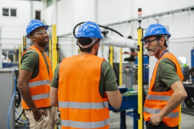 Güvenlik yeleği ve kask giyen çok ırklı endüstriyel mühendisler modern bir fabrikanın içindeki robot kolu denetliyor ve yenilikçi otomasyon çözümleri üzerinde tartışıyor ve işbirliği yapıyorlar.