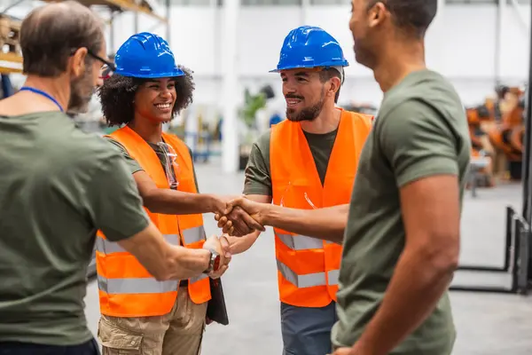 Güvenlik yelekleri ve şapka giyen çok ırklı erkek ve kadın mühendisler bir robotik fabrikasında el sıkışıyor, ileri teknoloji gelişiminde başarılı takım çalışmasını ve işbirliğini kutluyorlar.