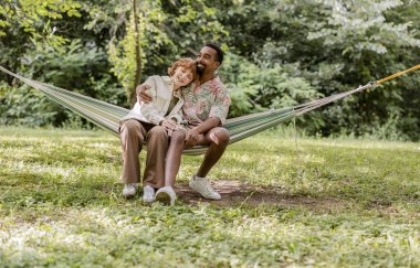 Mutlu çok ırklı çift, açık havada, yemyeşil bir hamakta birlikte, rahatlatıcı bir anı kucaklıyor ve eğleniyor.