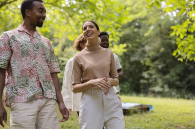 Farklı geçmişlere sahip neşeli arkadaşlar parkta yürüyor, konuşuyor, gülüyor, yaz toplantılarında bağlarını kutluyorlar.