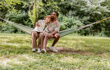 Mutlu çok ırklı çift, açık havada, yemyeşil bir hamakta birlikte, rahatlatıcı bir anı kucaklıyor ve eğleniyor.