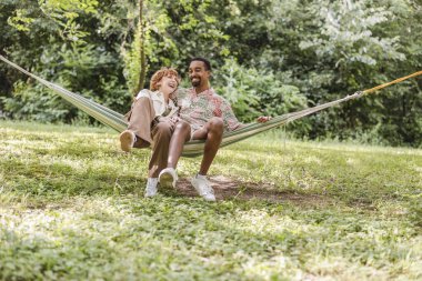 Mutlu çok ırklı çift hamakta birlikte gülüyor ve rahatlıyor. Yaz tatillerinin tadını çıkarıyorlar. Huzurlu doğal ortamlarında.