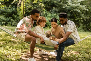 Dört neşeli çoklu etnik arkadaş parkta bir hamakta birlikte oturuyorlar, akıllı telefona bakıyorlar ve rahatlatıcı yaz tatili günlerinde birlikte vakit geçiriyorlar.