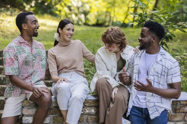 Four cheerful multi ethnic friends are sitting on a low brick wall in a park, laughing and chatting as they enjoy each other's company during a leisurely summer day clipart