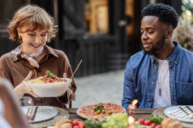 Genç bir kadın arkadaşlarıyla çok ırklı bir partide spagetti servis edip sıcak bir restoran bahçesinde yemek ve kahkaha atıyor.