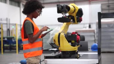 Kadın mühendis, modern bir fabrikada teknolojinin ve insan uzmanlığının endüstriyel otomasyon alanındaki entegrasyonunu gösteren bir robot kolu programlamak için bir tablet kullanıyor.