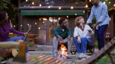 Çok ırklı arkadaşlar, dinlendirici bir tatil akşamında ahşap bir dağ evinin önünde ellerini ısıtıyorlar. Sıcaklığın ve arkadaşlığın tadını çıkarıyorlar.