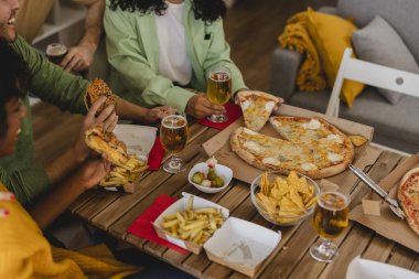 Bir grup arkadaş yemeğin tadını çıkarıyor, pizza, sandviç, patates kızartması, nacho ve bira paylaşıyor, ahşap bir masada oturuyor, eğleniyor ve sosyalleşiyorlar.