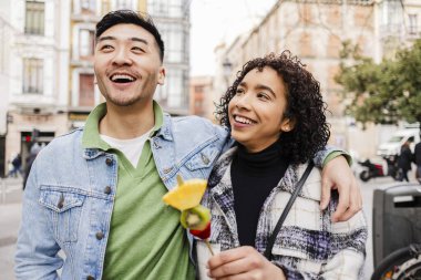 Genç, çok kültürlü bir çift gülümsüyor ve kol kola yürüyorlar. Neşeli bir şehirde güneşli bir sevgililer gününde lezzetli bir meyve şişin tadını çıkarıyorlar. Neşe ve sevgi saçıyorlar.