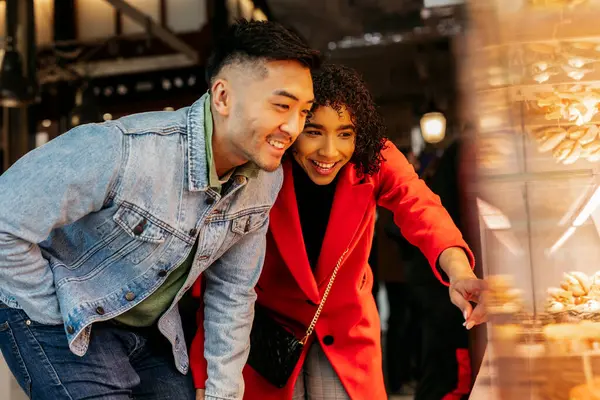 Neşeli, genç, çok kültürlü bir çift gülümseyip işaret ederken pastanenin vitrininde lezzetli hamur işleri seçiyor Sevgililer Günü 'nü tatlı ikramlarla ve mutlu anlarla kutluyorlar.