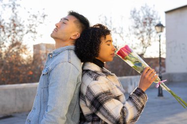 Young multiracial couple is standing back to back outdoors, enjoying a moment of peace and tranquility as the woman smells a single red rose, symbolizing love and connection on valentine's day clipart