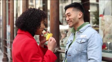 Genç bir kadın, erkek arkadaşına vitrinin önünde dikilirken bir meyve şişi sunuyor. Çift canlı şehir caddesinde birlikte neşeli bir anın tadını çıkarıyor.