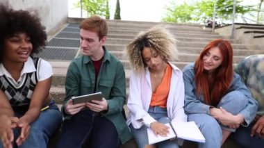 Dört neşeli çoklu etnik gruptan oluşan bir grup kampüs merdivenlerinde oturmuş, canlı bir açık hava ortamında birbirlerinin arkadaşlığını çalışırken ve eğlenirken dijital tabletle uğraşıyorlar.