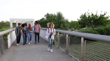 Bir grup genç, köprüde yalnız bir öğrencinin arkasında yürürken gülüyor ve işaret ediyor, akran baskısını vurguluyor ve okul ortamında zorbalık yapıyor.