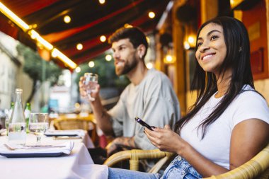 Lizbon, Portekiz 'deki bir açık hava restoranında erkek arkadaşıyla yemek yerken gülümseyen ve cep telefonu kullanan genç bir kadın yaz akşamının tadını çıkarıyor.