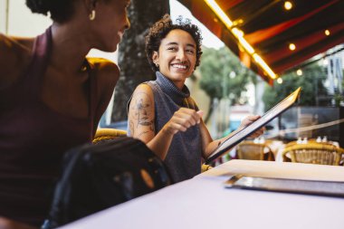 İki neşeli bayan arkadaş canlı bir Lizbon restoranında bir masada oturmuş menüyü okurken ve yemek siparişleri sırasında gülümserken yaz güneşinin tadını çıkarıyorlar.