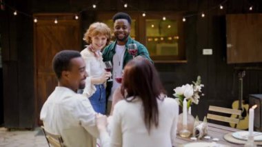 Bir grup farklı arkadaş kırmızı şarap kadehlerini kaldırıyor, açık hava partisinde kutlama yapıyorlar, etrafları tel ışıkları ve neşeli atmosferle çevrili.