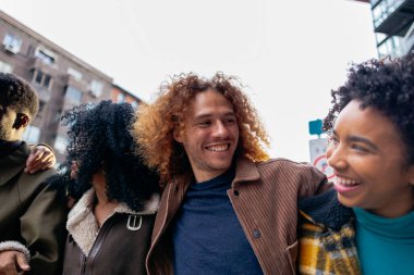 Bir grup neşeli, etnik çeşitlilikte yetişkin öğrenci şehirde birlikte yürüyor, birbirlerine sarılıyor ve gülümsüyor, üniversiteden sonra boş zamanlarının tadını çıkarıyorlar.