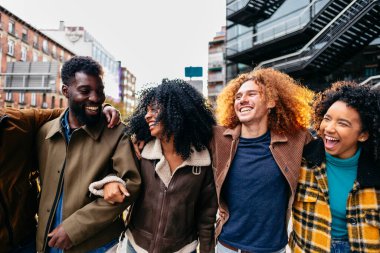 Bir grup neşeli, çoklu etnik arkadaş şehirde birlikte yürüyor, birbirlerini kucaklıyor ve dışarıda şehir ortamında eğlenirken aynı zamanda kahkahalarını paylaşıyorlar.