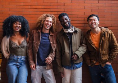 Neşeli bir grup genç arkadaş bir tuğla duvarın önünde durmuş, kahkahalarını paylaşıyor ve birlikte eğleniyorlar. Görüntü arkadaşlığı, çeşitliliği ve pozitif enerjiyi aktarır.