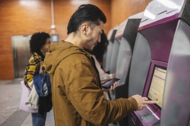 Asyalı turist bir metro istasyonundaki self servis makinesinden tren bileti alırken, diğer insanlar kuyrukta beklerken akıllı telefon uygulaması ile işlemi tamamlıyor.
