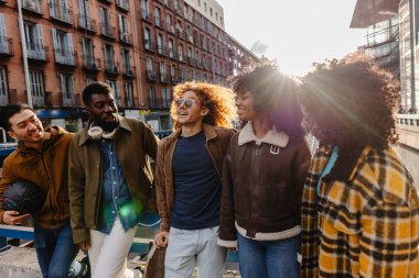 Güneşli bir şehirde birlikte yürüyüp konuşan beş neşeli çoklu etnik arkadaş, birbirlerinin arkadaşlığından keyif alıyorlar ve çevrelerindeki canlı şehir atmosferini kucaklıyorlar.