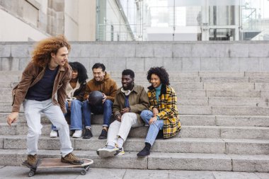 Bir grup çok ırklı arkadaş dışarıda eğleniyor, gülüyor ve sohbet ediyorlar. Bu arada genç bir kaykaycı, canlı bir şehirde şehir basamaklarında dinleniyor.