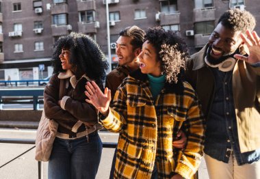 Dört neşeli çoklu etnik arkadaş şehirde yürürken, bir yandan gülümserken, bir yandan da güneşli bir günün tadını çıkarırken bir yandan da el hareketleriyle birini selamlıyorlar.