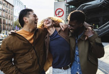 Üç neşeli genç adam, modern bir şehir ortamında birbirlerine sarılıp gülüyor ve eğleniyorlar.