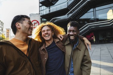 Güneşli bir günde, modern bir binanın yanında birlikte gülüp eğlenen üç neşeli genç adam var.